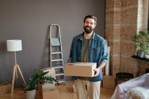 Horizontal shot of cheerful young man with beard on face wearing casual clothes holding box standing in his new apartment